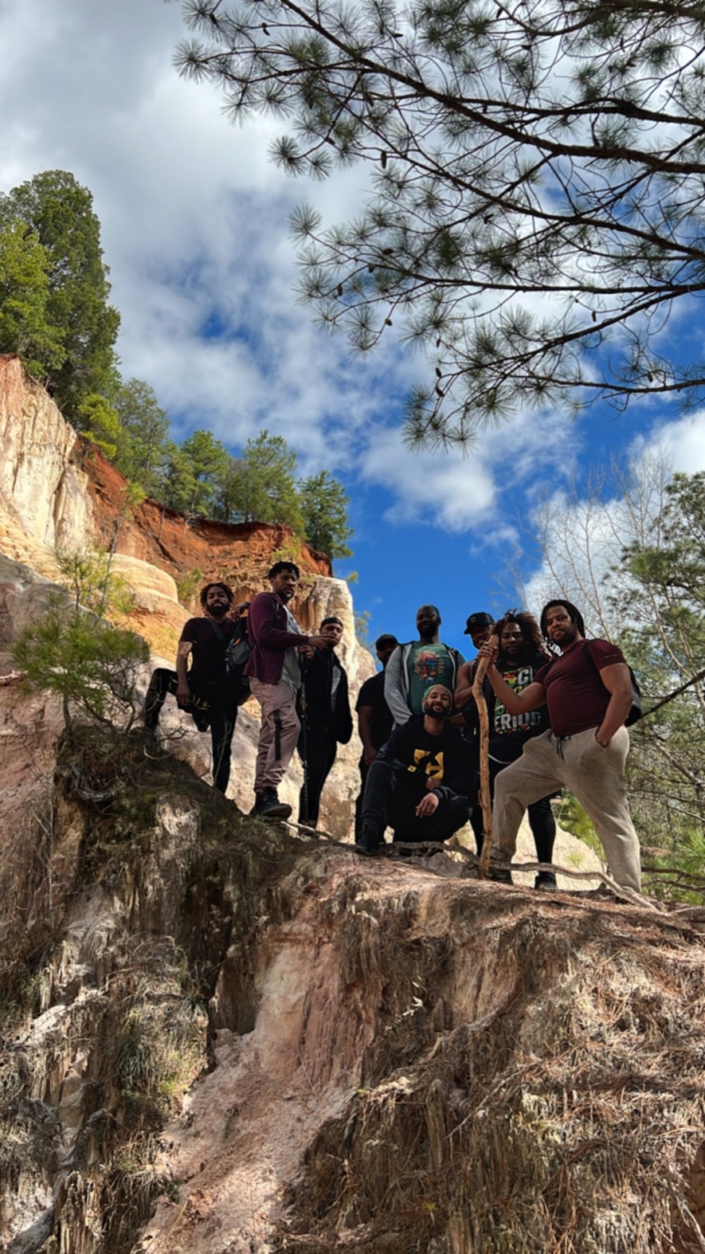 A group of Black men pause on a steep rock ledge beneath blue sky and pine branches.